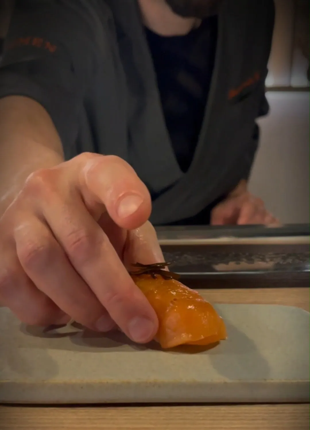 La mano dello chef posa un nigiri di salmone su un piatto di gres, sul banco di legno.